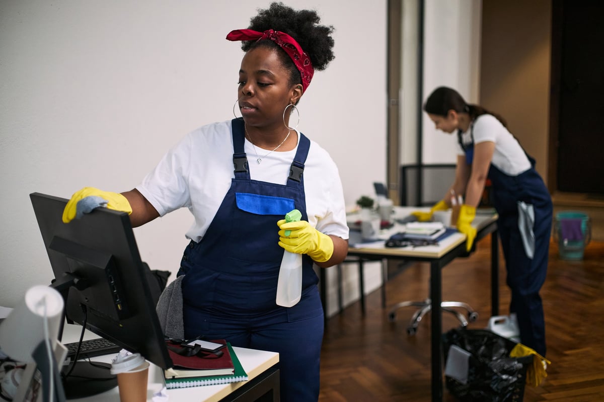 Professional cleaners in modern office — one cleaning computer monitor while another organizes desk