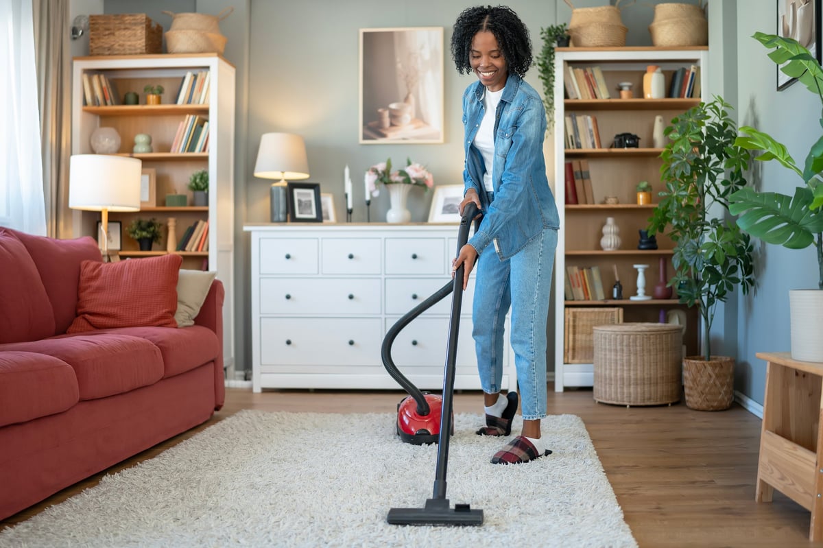 Smiling cleaner vacuuming a plush white carpet in a bright modern living room — recurring cleaning keeps your home always guest-ready